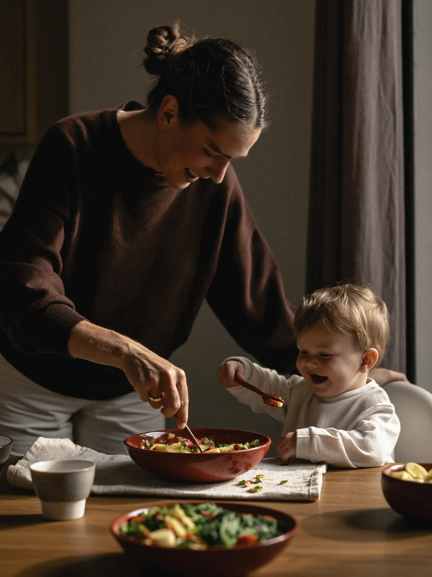 Mother feeding her toddler a colourful meal at a kitchen table, both laughing